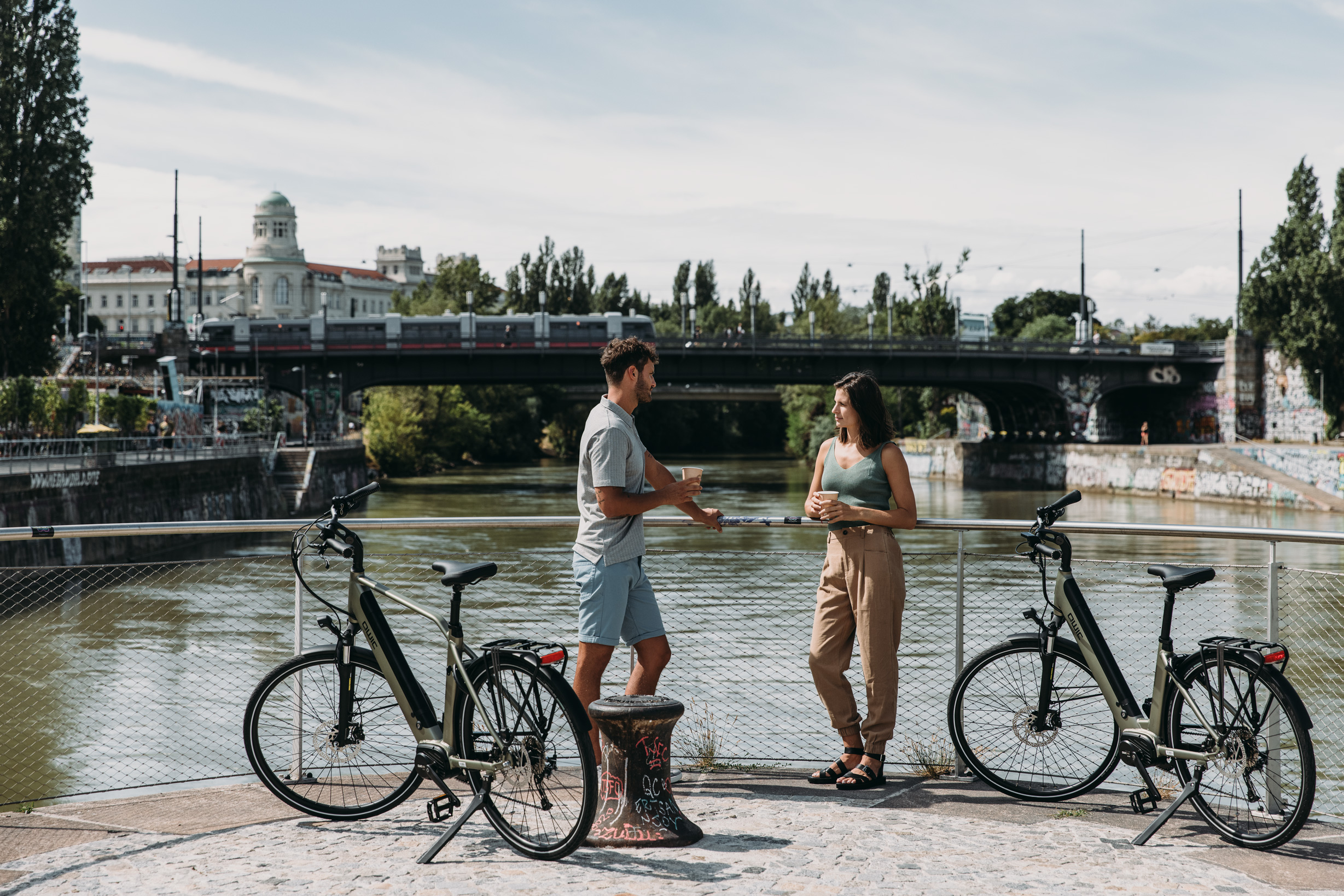 2 Personen stehen mit ihren Fahrrädern am Wasser. Im Hintergrund die Urania